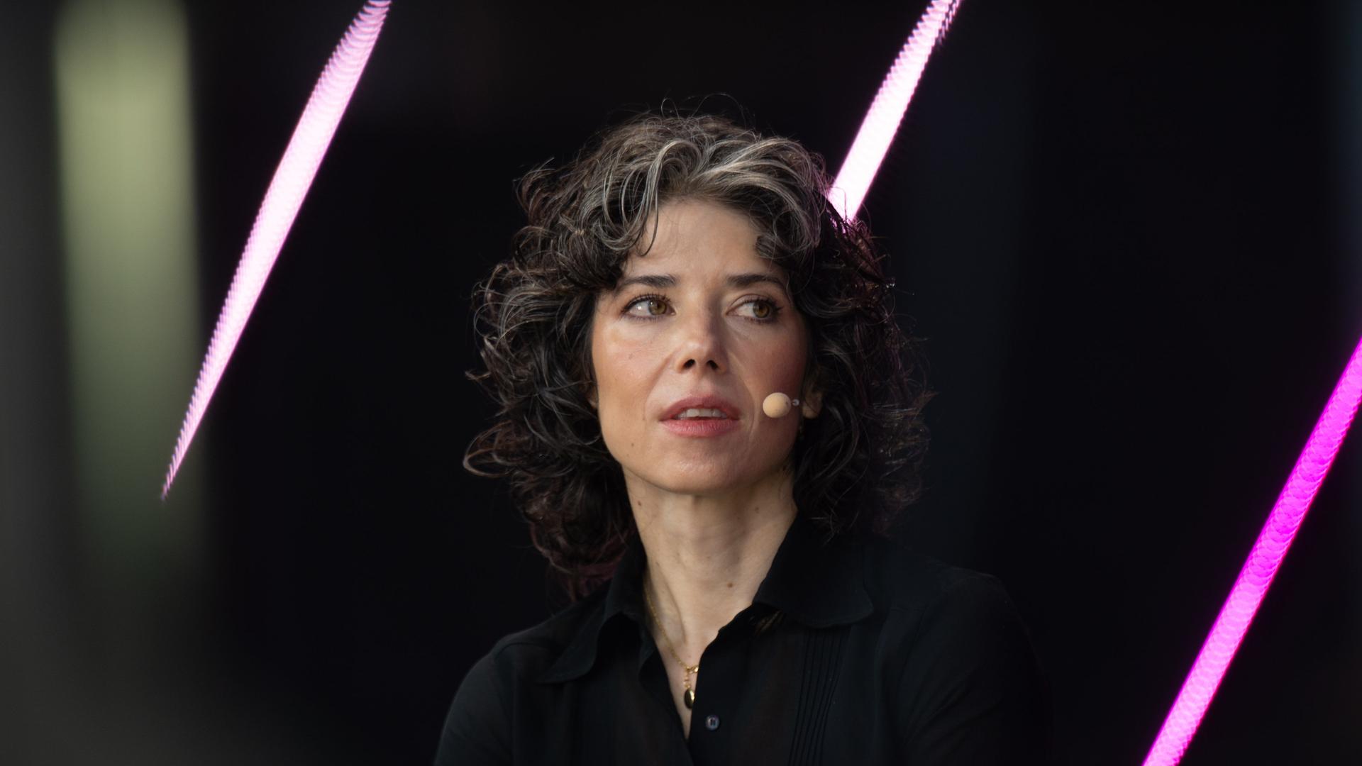 Meredith Whittaker sitzt auf einer schwarzen Bühne, die von schrägen, pinken Lichtstreifen im Hintergrund unterbrochen wird. Sie trägt ein schwarzes Oberteil und ein Bügelmikrofon und schaut zur Seite.