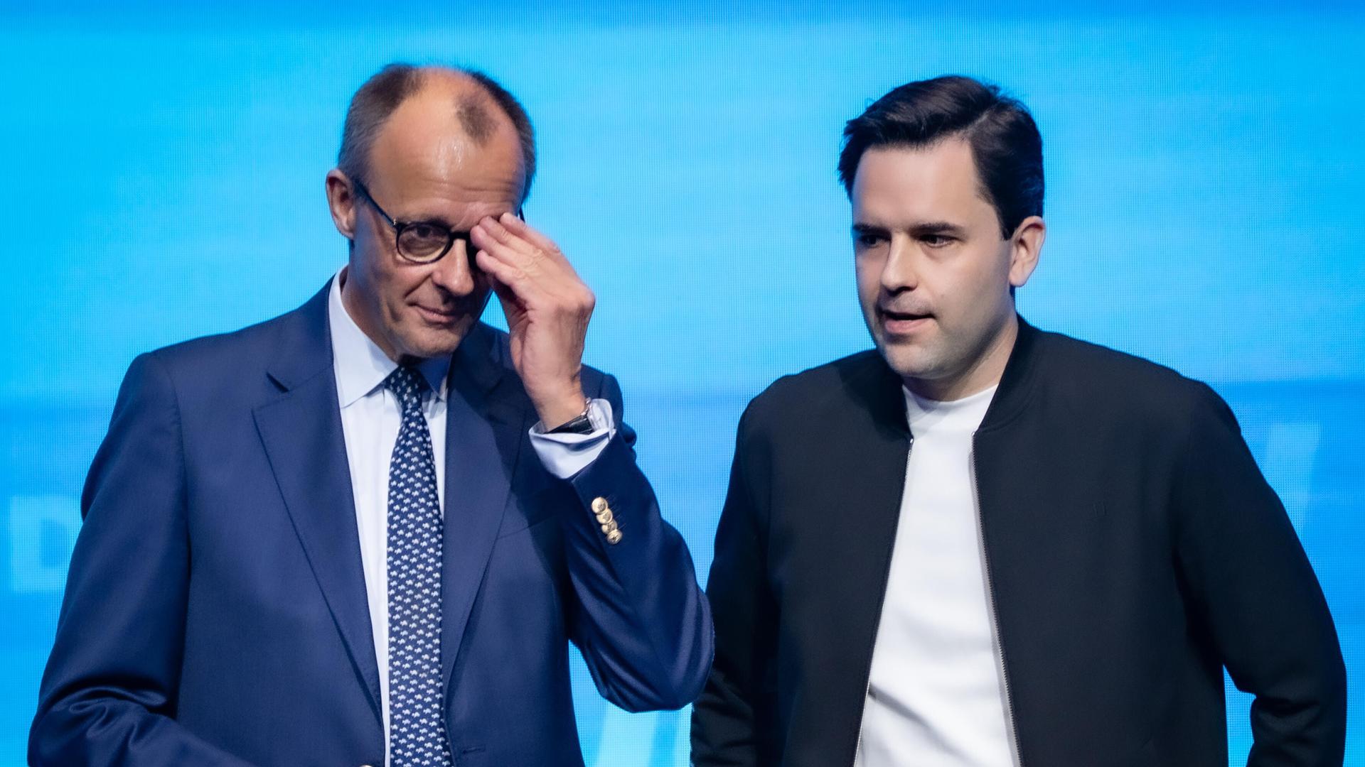 Bundeskanzler Friedrich Merz steht neben Johannes Winkel, Bundesvorsitzender Jungen Union, und fasst sich an die Brille, beide sehen nicht sehr glücklich aus