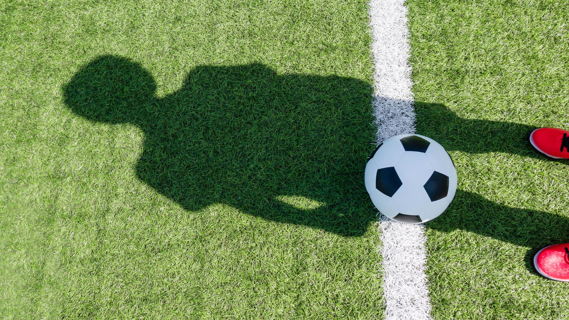 Auf einem Fussballfeld liegt ein Fussball und eine Person mit roten Schuhen wirft einen Schatten auf das Feld und den Ball.