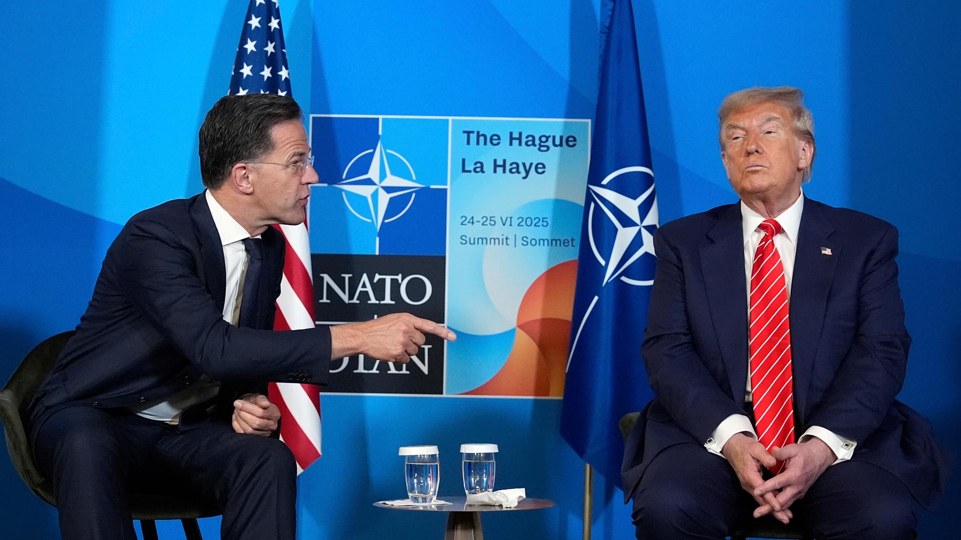 Präsident Donald Trump und NATO-Generalsekretär Mark Rutte unterhalten sich bei einer Pressekonferenz im Rahmen des NATO-Gipfels in Den Haag. Mark Rutte wendet sich Donald Trump mit ausgstrecktem Zeigefinger zu. Im Hintergrund sieht man die NATO-Flagge.