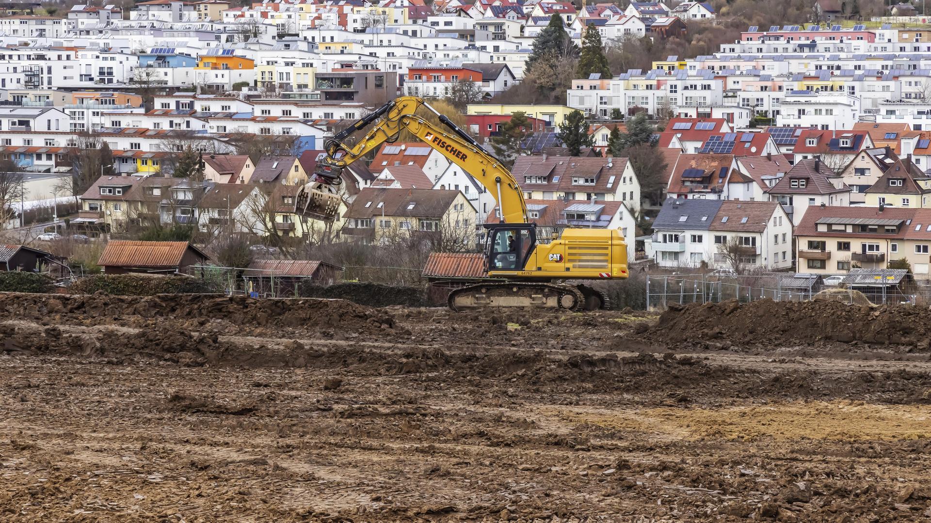 Ein Bagger steht auf Ackerland. Im Hintergrund sieht man mehrere Häuser. 