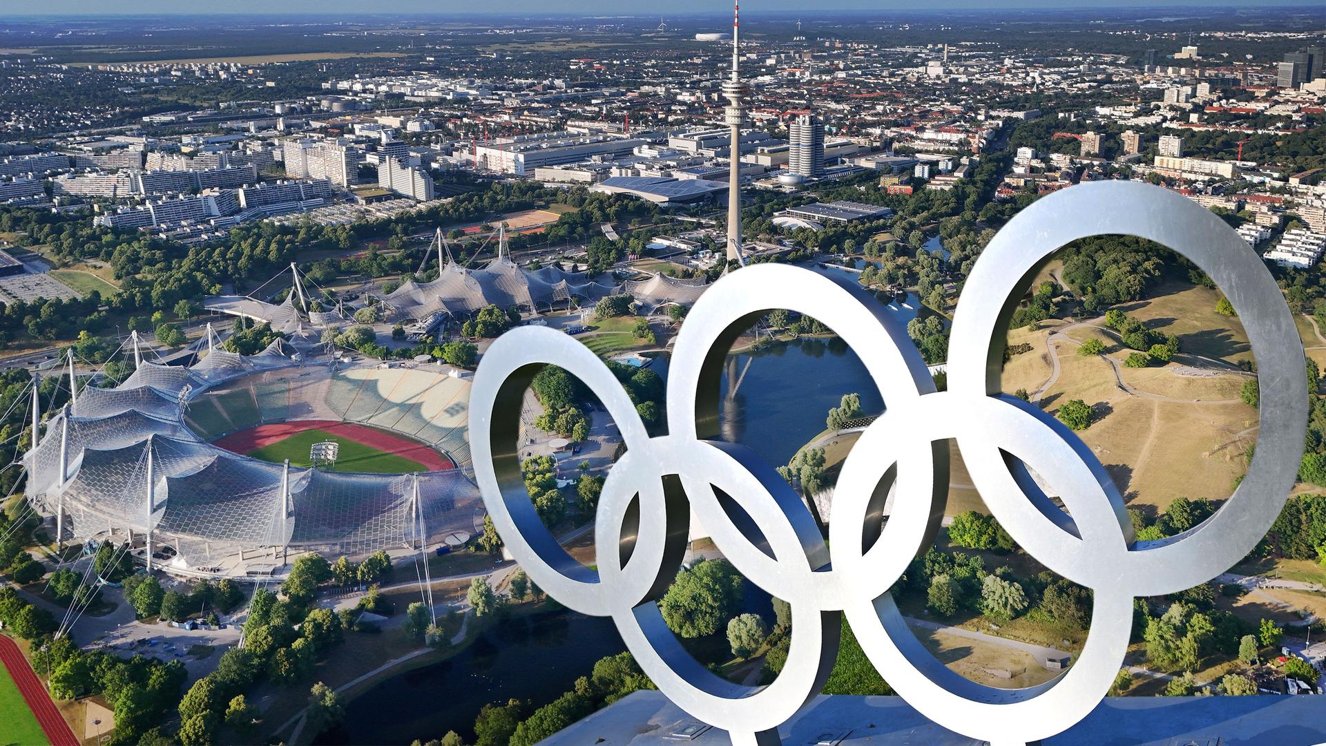 Luftaufnahme des Olympiaparks mit Olympiastadion, Olympia-Schwimmhalle und Olympiahalle, davor die Olympischen Ringe auf einer Dachkonstruktion (Fotomontage) 