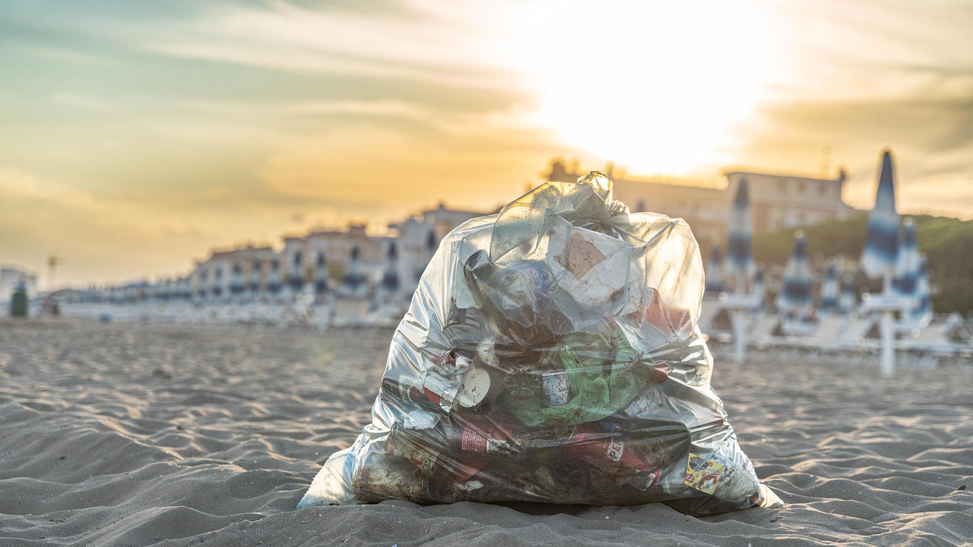 Ein gefüllter Müllsack mit Plastikabfällen liegt im Sand an einem Strand.