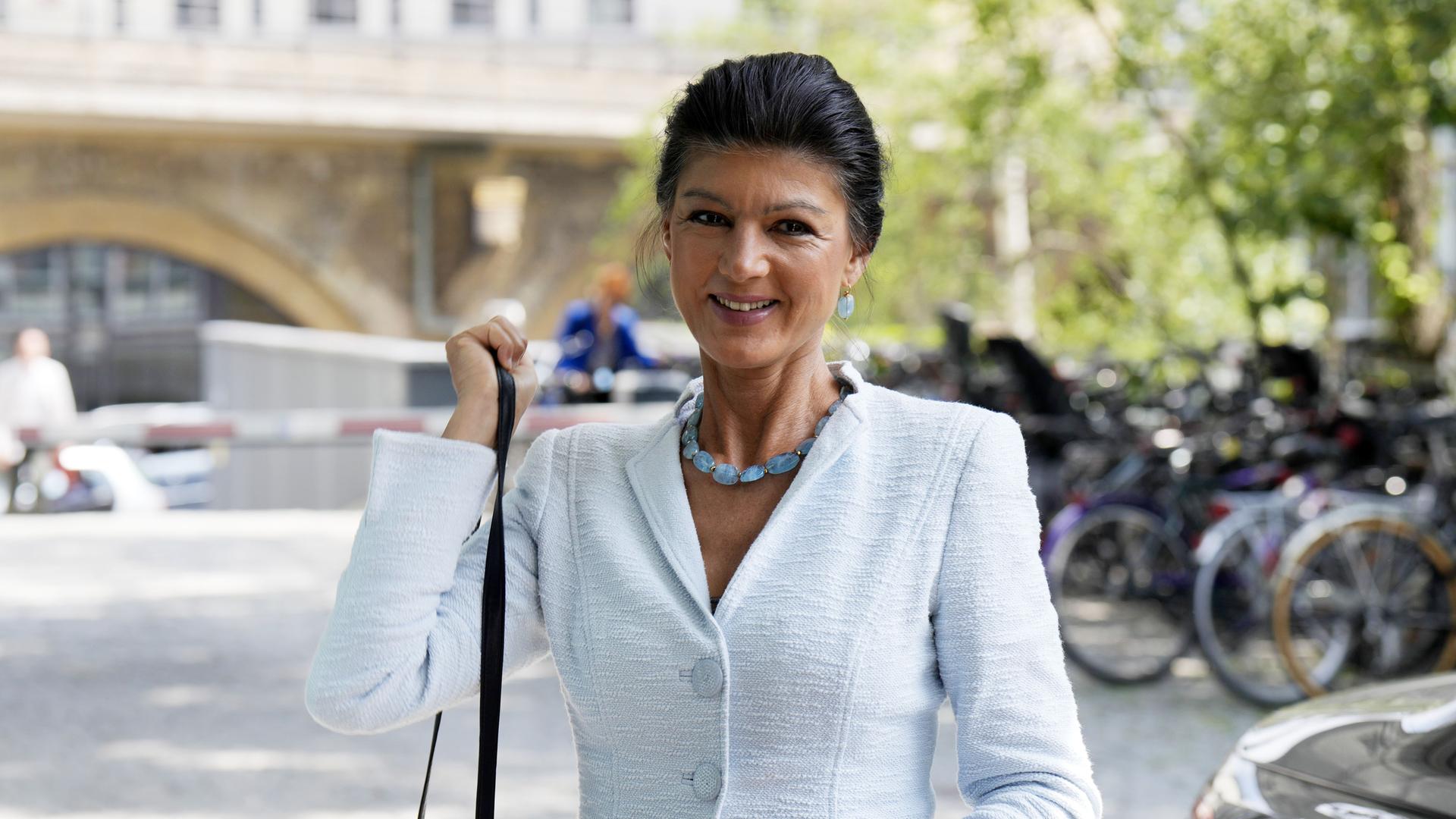 Sahra Wagenknecht lächelt in die Kamera und hängt sich dabei eine Handtasche an die Schulter. Im Hintergrund sieht man verschwommen abgestellte Fahrräder.