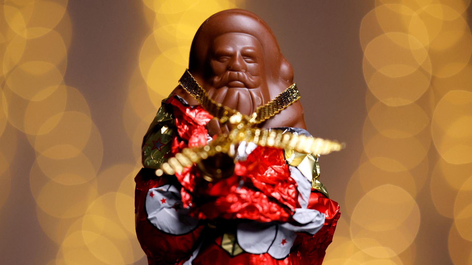 Ein teilweise ausgepackter Schokoladen-Weihnachtsmann in rot-goldener Folie. Der Hintergrund ist unscharf und besteht aus warmen, goldenen Lichtkreisen. (Symbolbild) 