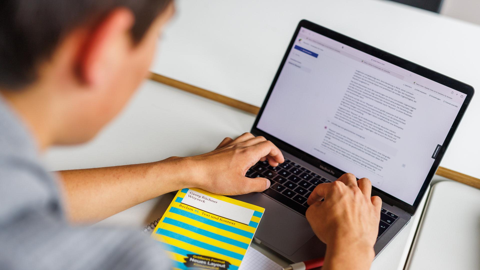 Ein Schüler sitzt vor einem Laptop und benutzt Chat-GPT. Vor ihm liegt ein gelbes Buch: "Woyzeck" von Büchner.