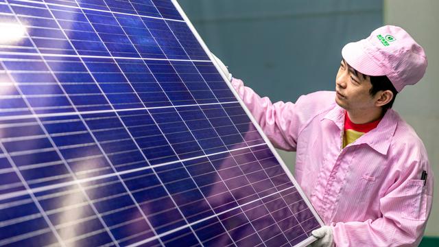 Ein chinesischer Mann hält ein großes Solarmodul in den Händen und schaut prüfend darauf. Er trägt einen rosafarbenen Arbeitsoverall und Mütze. 