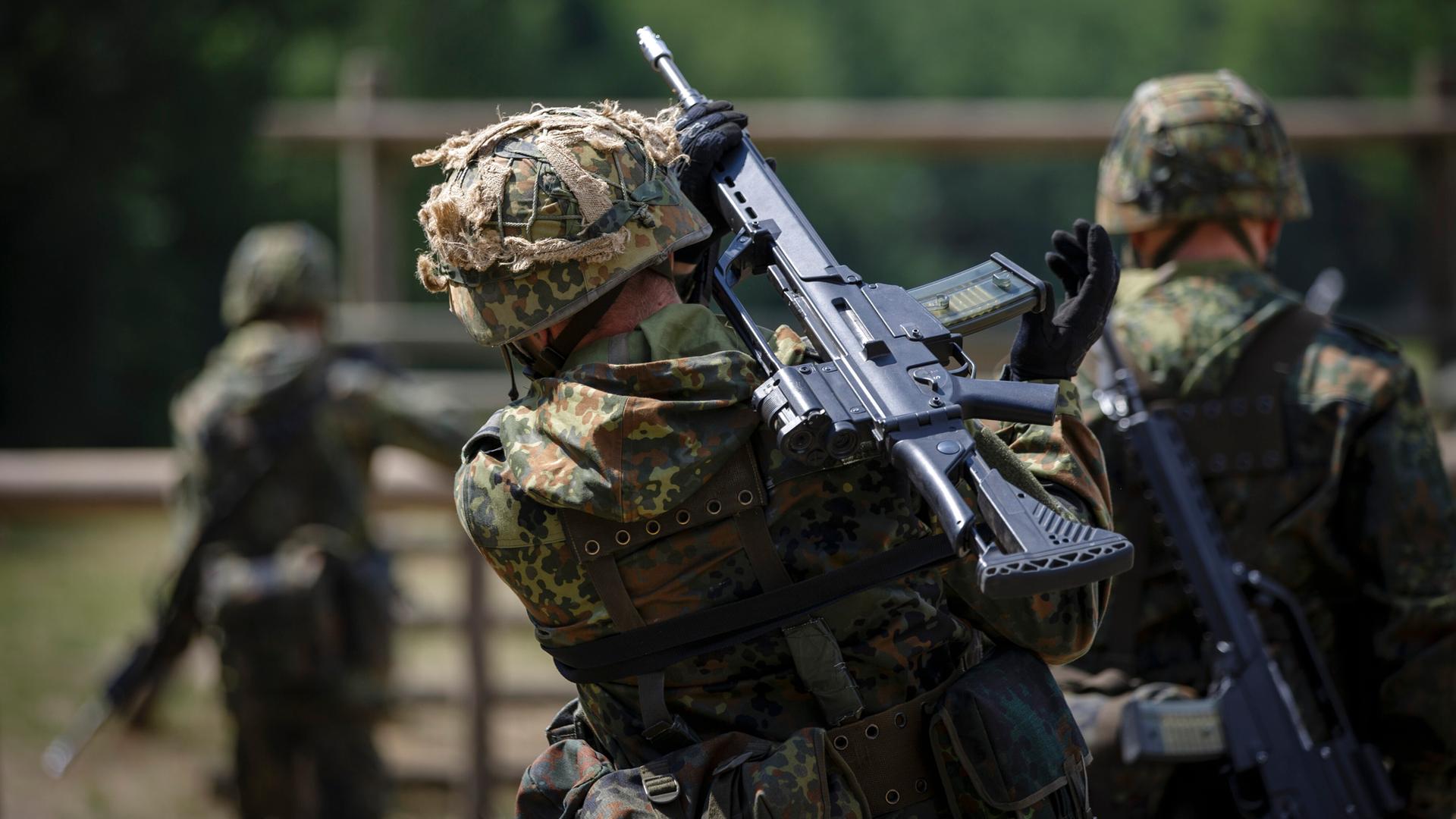 Drei Rekruten marschieren auf einem Gelände, sie haben Gewehre geschultert. Alle drei tragen Tarnanzüge und sind von hinten zu sehen.