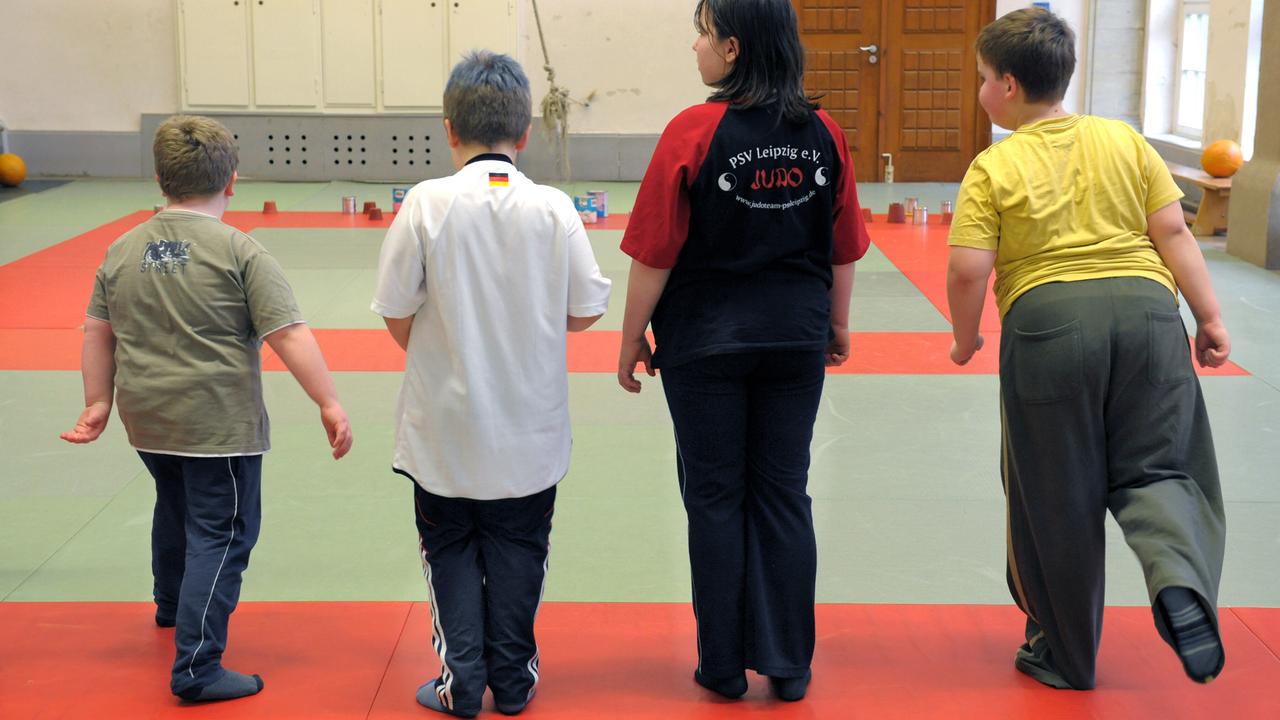 Vier Kinder mit Übergewicht stehen in einer Turnhalle auf einer Matte mit dem Rücken zum Betrachter. Ein Junge rechts hebt das rechte Bein nach hinten. 