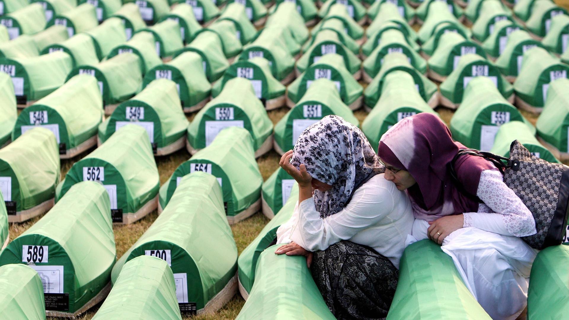 Zwei Frauen trauern während der Beerdigung von neu identifizierten bosnischen Muslimen in Srebrenica 2010. Sie sitzen zwischen hunderten nummerierten Särgen, die mit grünem Stoff bespannt sind.