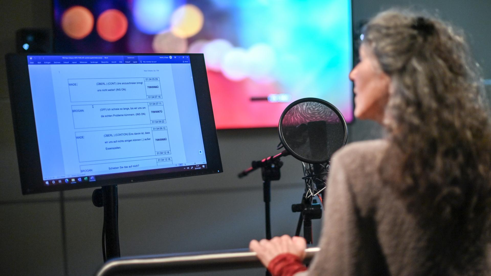 Eine Frau steht in einem Synchronstudio hinter einem Mikrofon und einem Bildschirm, der Dialoge anzeigt.