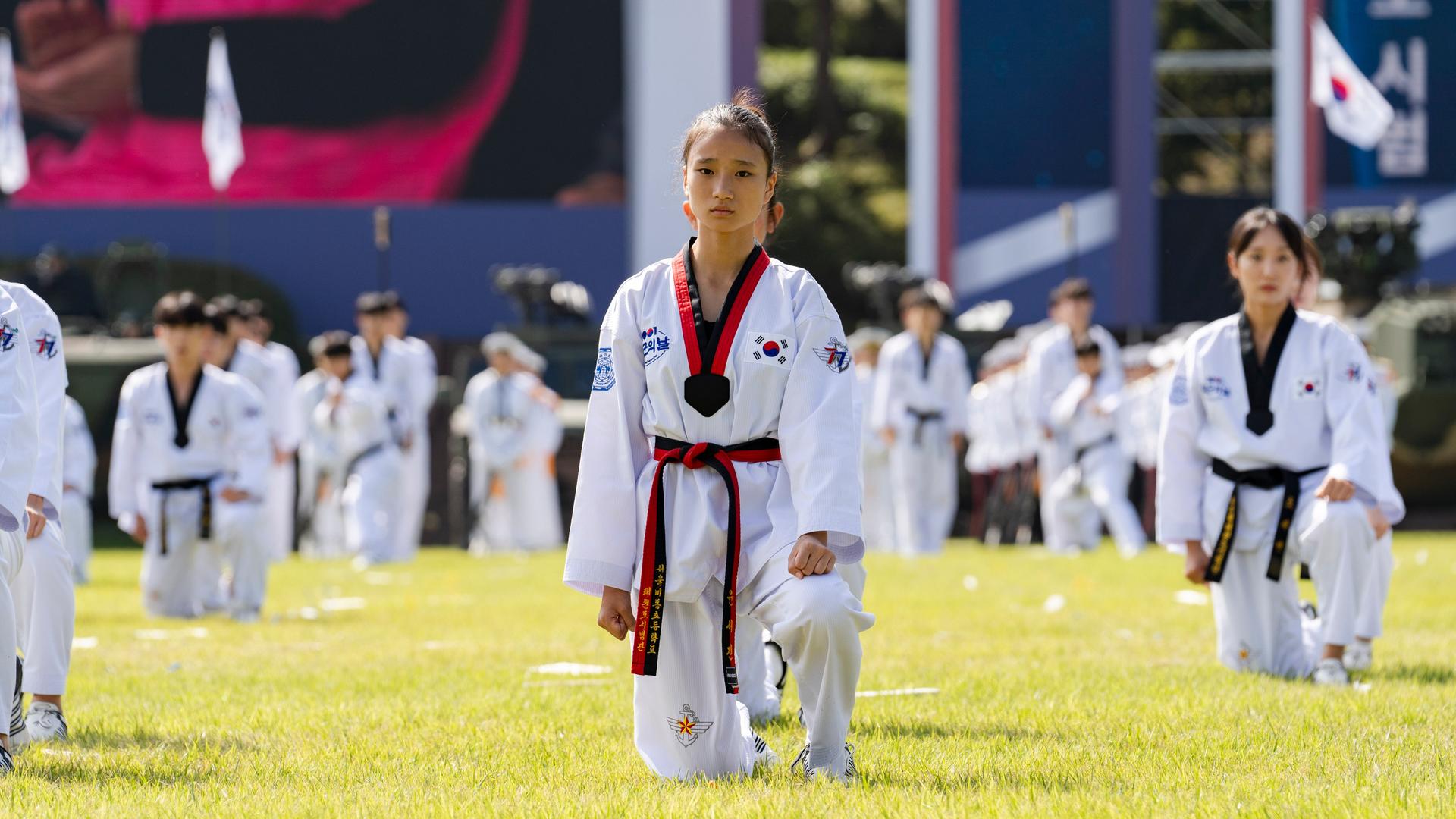 Bei einer großen Veranstaltung unter freiem Himmel sind Taekwondo-Kämpfer aufgestellt. Eine junge Frau in ihrem weißen Kampfanzug schaut in die Kamera.