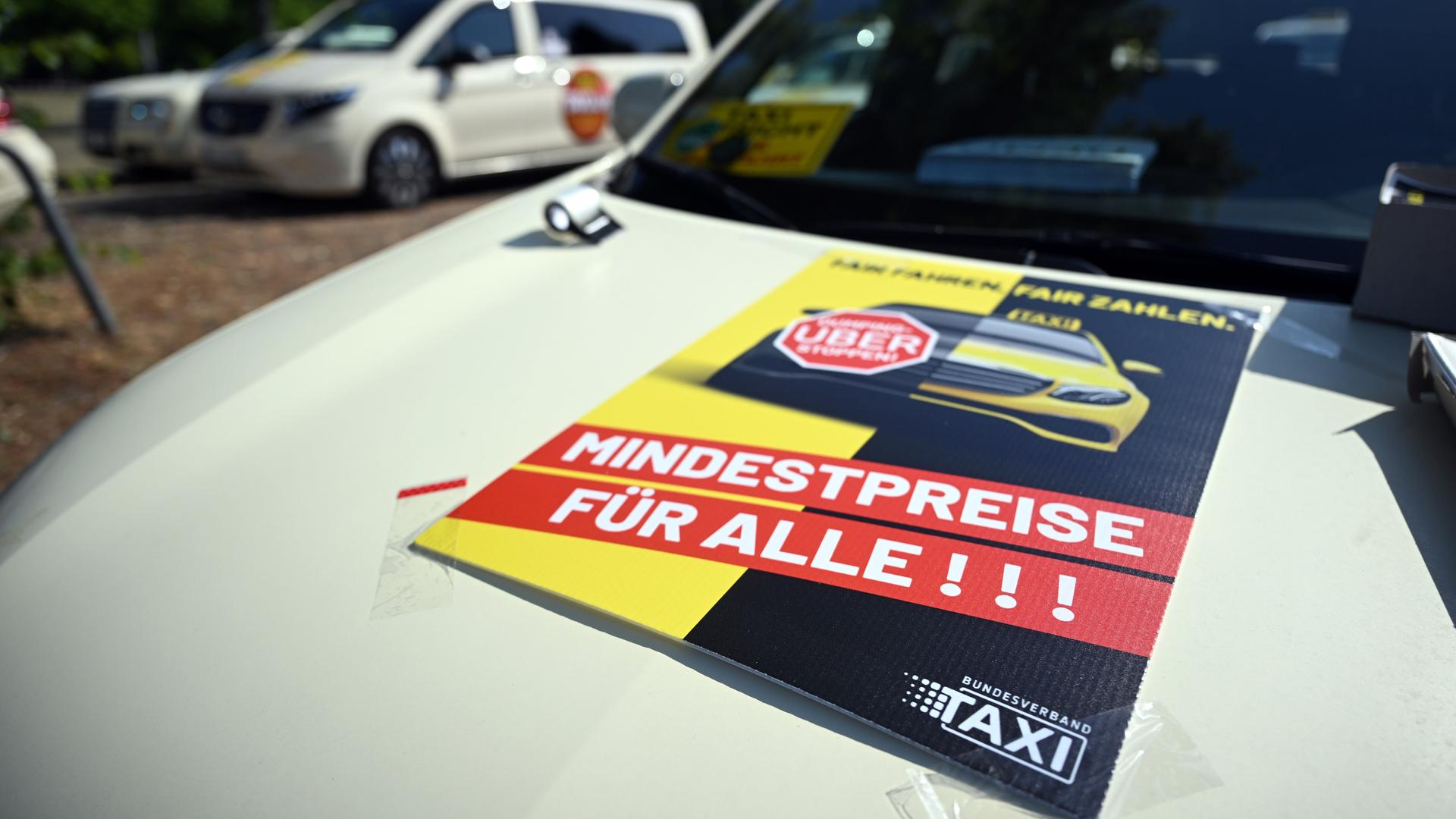 Taxis stehen auf einem Messe-Parkplatz um sich zu einem Autokorso zu sammeln. Auf einer Motorhaube steht auf einem Plakat "Fair zahlen. Mindestpreise für alle"