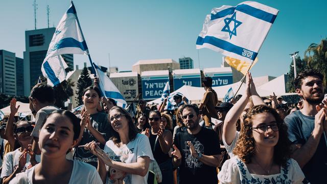 Menschen auf dem Platz der Geiseln feiern die Rückkehr der letzten lebenden Geiseln aus der Gewalt der Hamas. Über ihnen sind Israel-Flaggen zu sehen.