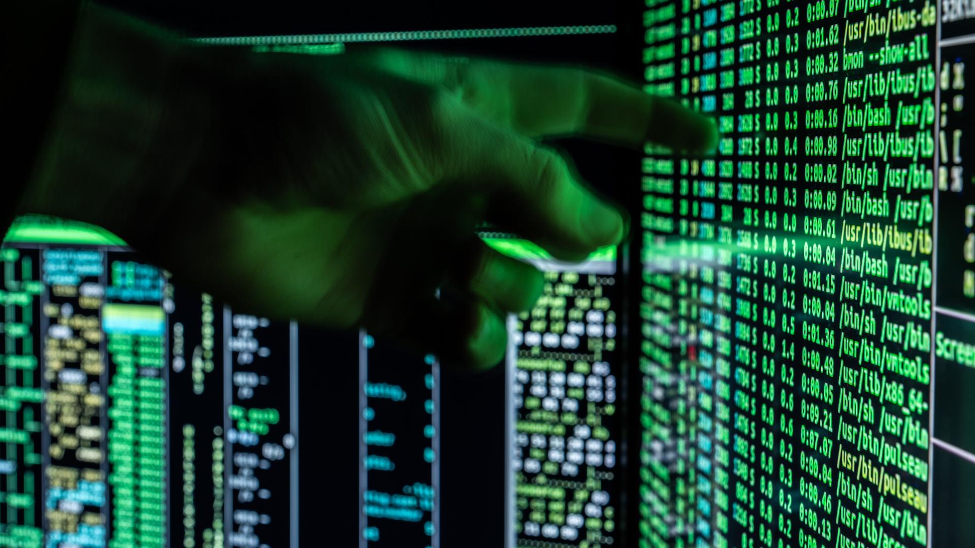 Ein Mann hält seine Hand vor ein fiktives Hacker-Programm, das auf zwei Bildschirmen eines Computers zu sehen ist