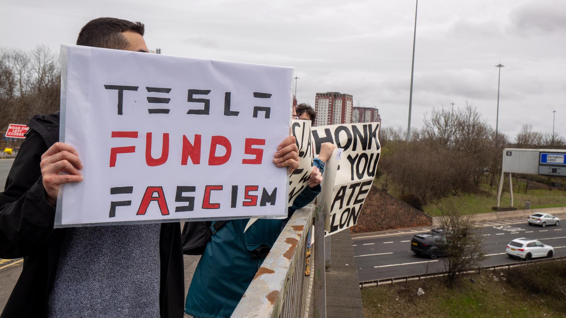 Demonstranten halten Schilder auf einer Brücke über einer Straße hoch, darauf steht: "Tesla funds fascism" oder "honk if you hate Elon".