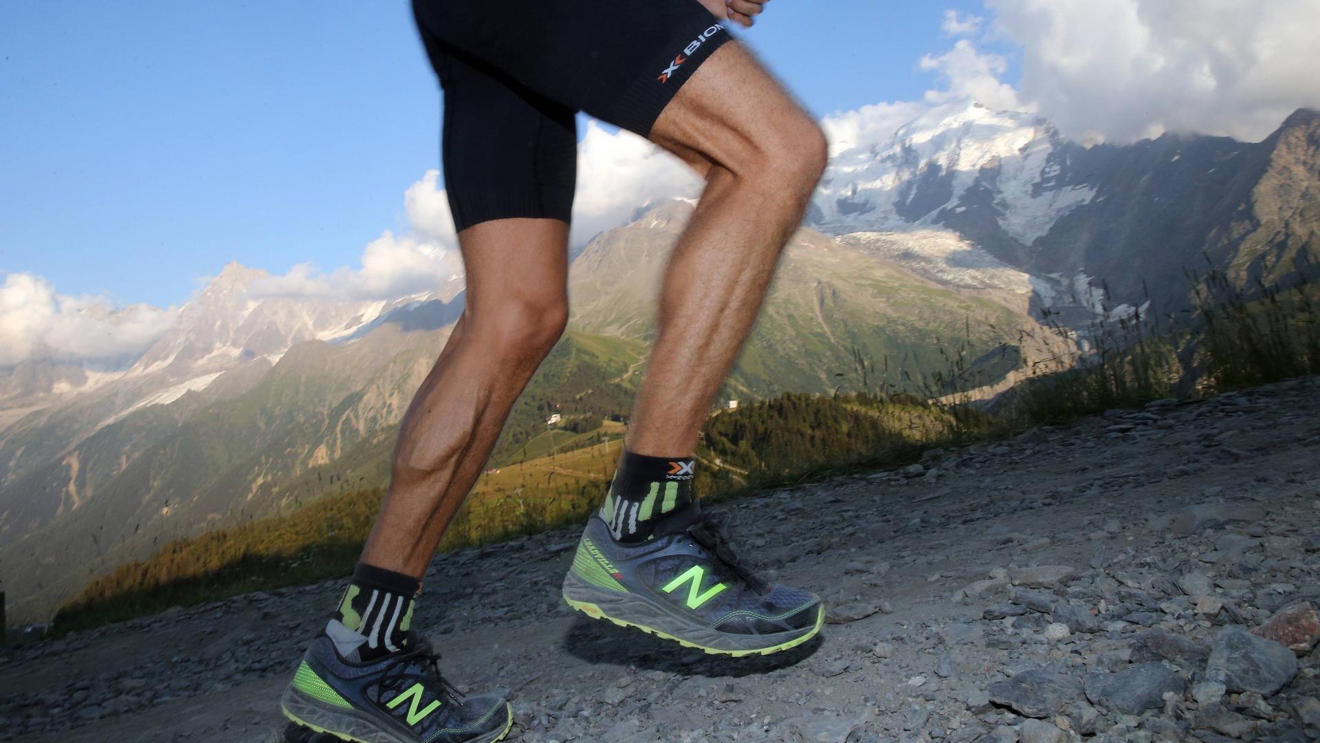 Nahaufnahme der Beine eines Läufers in professioneller Ausrüstung, der einen Pfad in den Alpen bergauf läuft.
