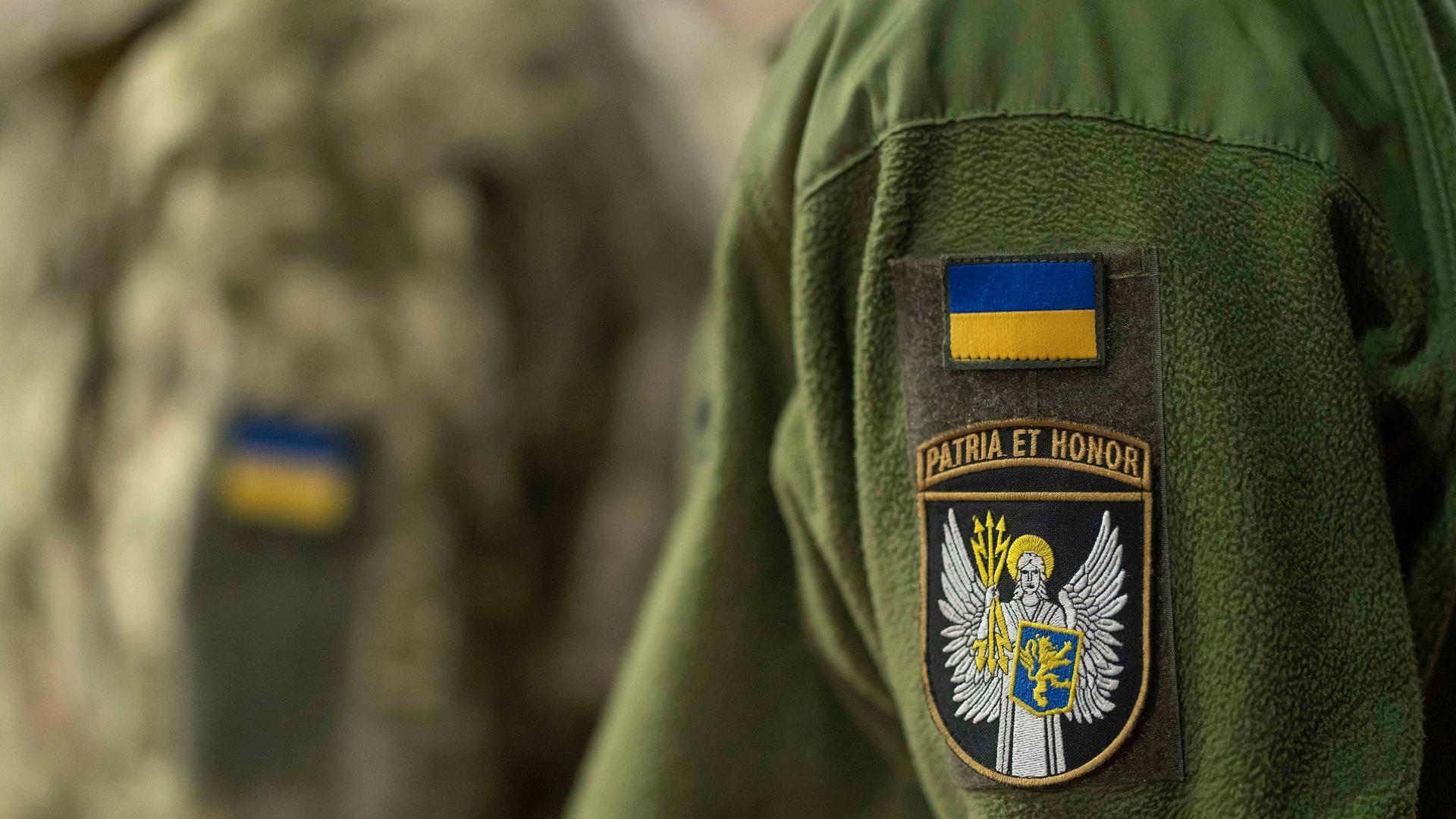 Zwei Personen tragen eine grünfarbene Uniform der ukrainischen Armee. Nur die Jacken sind zu sehen. Auf beiden ist je ein Aufnäher mit der ukrainischen Flagge genäht.