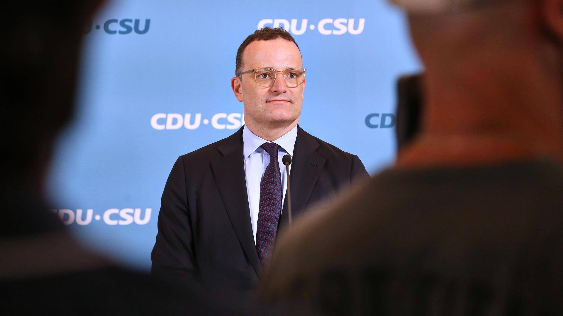 Jens Spahn steht vor blauem Hintergrund mit CDU/CSU-Logo vor einem Mikrofon. Am Rand sieht man zudem die Umrisse von anderen Personen
