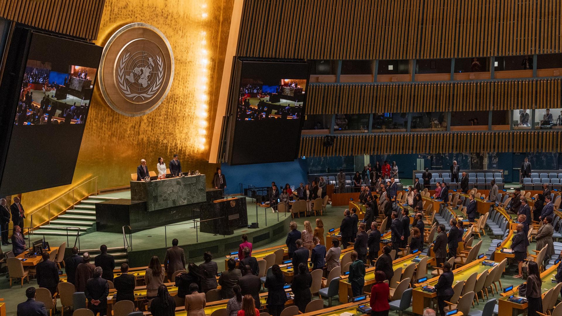 Blick in den Plenarsaal der UNO in New York, wo die Delegierten stehen. An der goldfarbenen Rückwand sieht man das Emblem der Organisation.