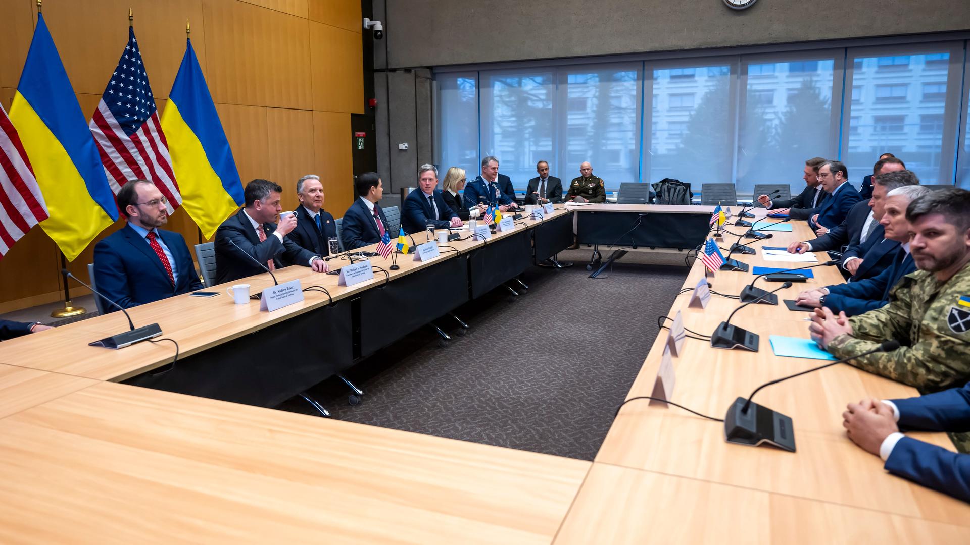 Menschen sitzen sich an einem langen Tisch gegenüber. Sie kommen aus den Ländern USA und Ukraine und verhandeln über einen Friedens-Plan für die Ukraine. 