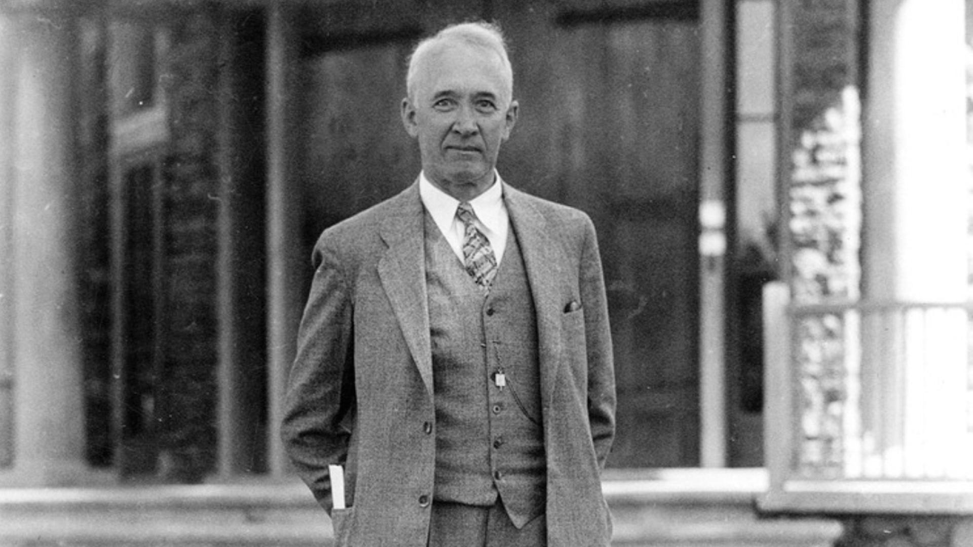 Ein Schwarz-Weiß-Bild zeigt den Astronomen Vesto Melvin Slipher, der in einem grauen Anzug mit Weste und Krawatte vor einem Gebäude für ein Foto posiert.