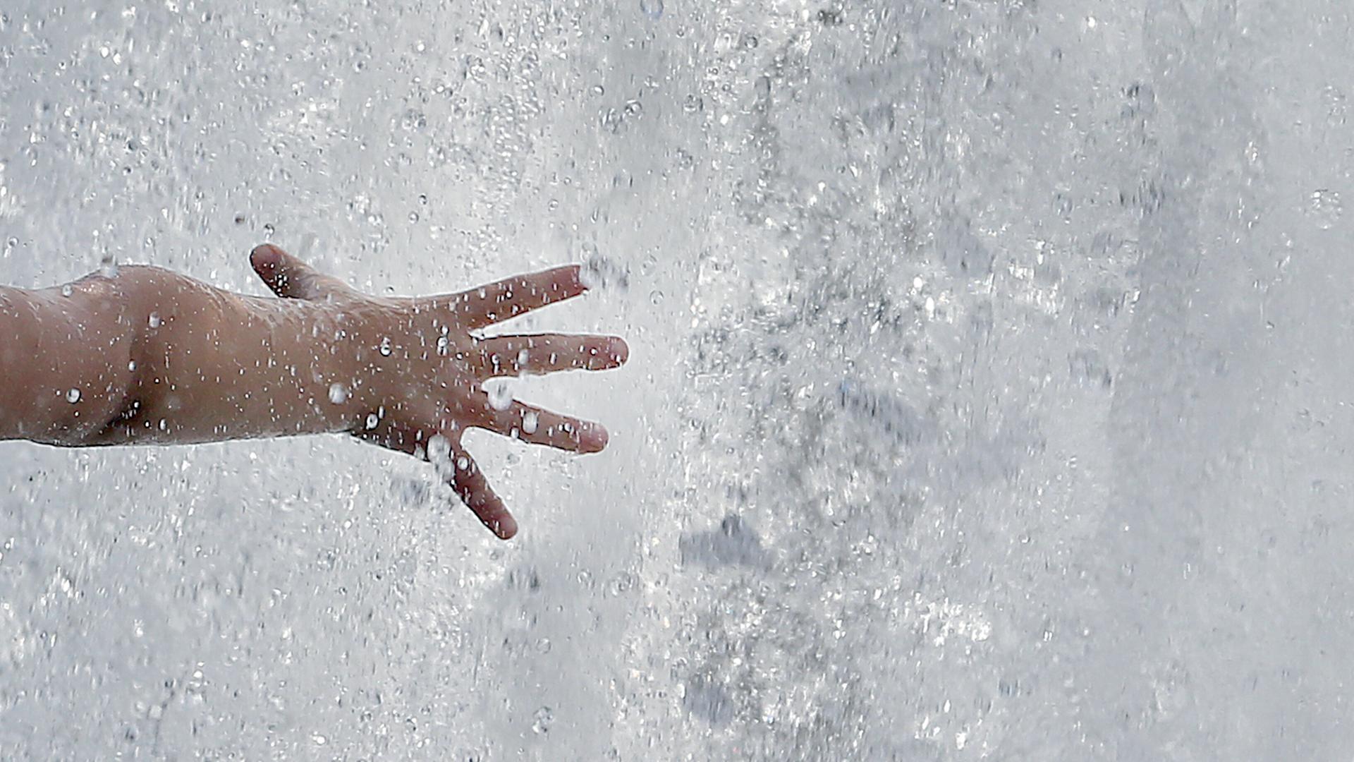 Das Bild zeigt eine Kinderhand, die bei bei Temperaturen um 31 Grad Celsius in eine Springbrunnenanlage mit sprudelndesm Wasser greift.