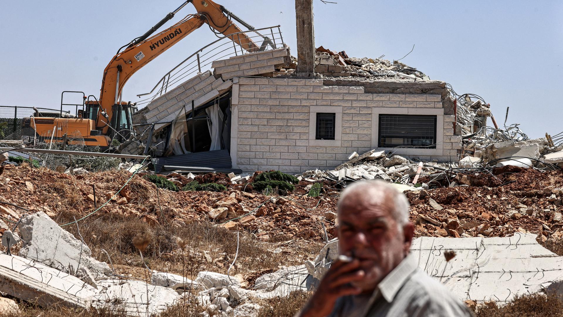 Ein Mann raucht eine Zigarette, während israelische Bagger am 4. August 2025 im Dorf Judeira südlich von Ramallah im besetzten Westjordanland ein Gebäude abreißen. 