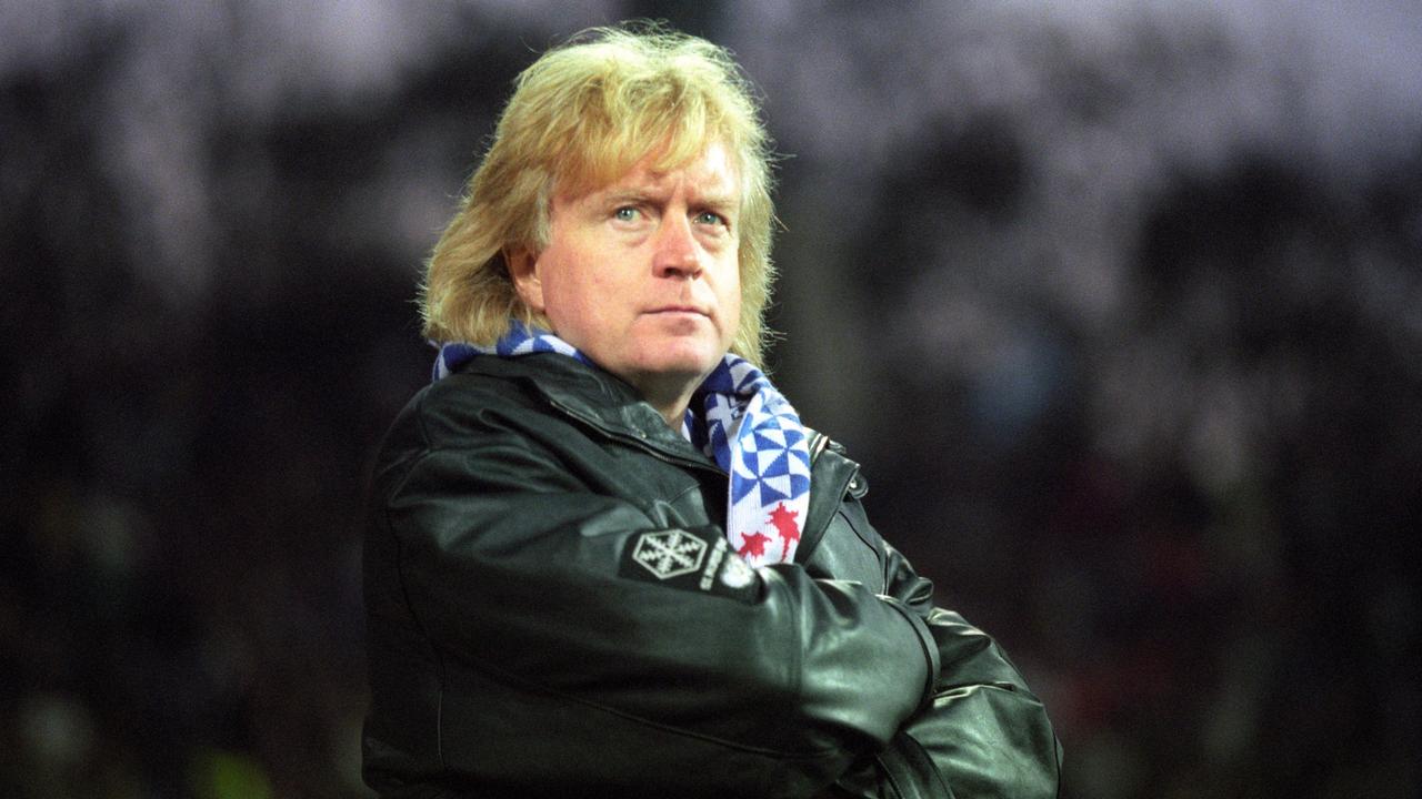 Winnie Schäfer steht mit verschränkten Armen auf dem Platz. Er trägt den blauen KSC-Schal zu einer schwarzen Lederjacke.